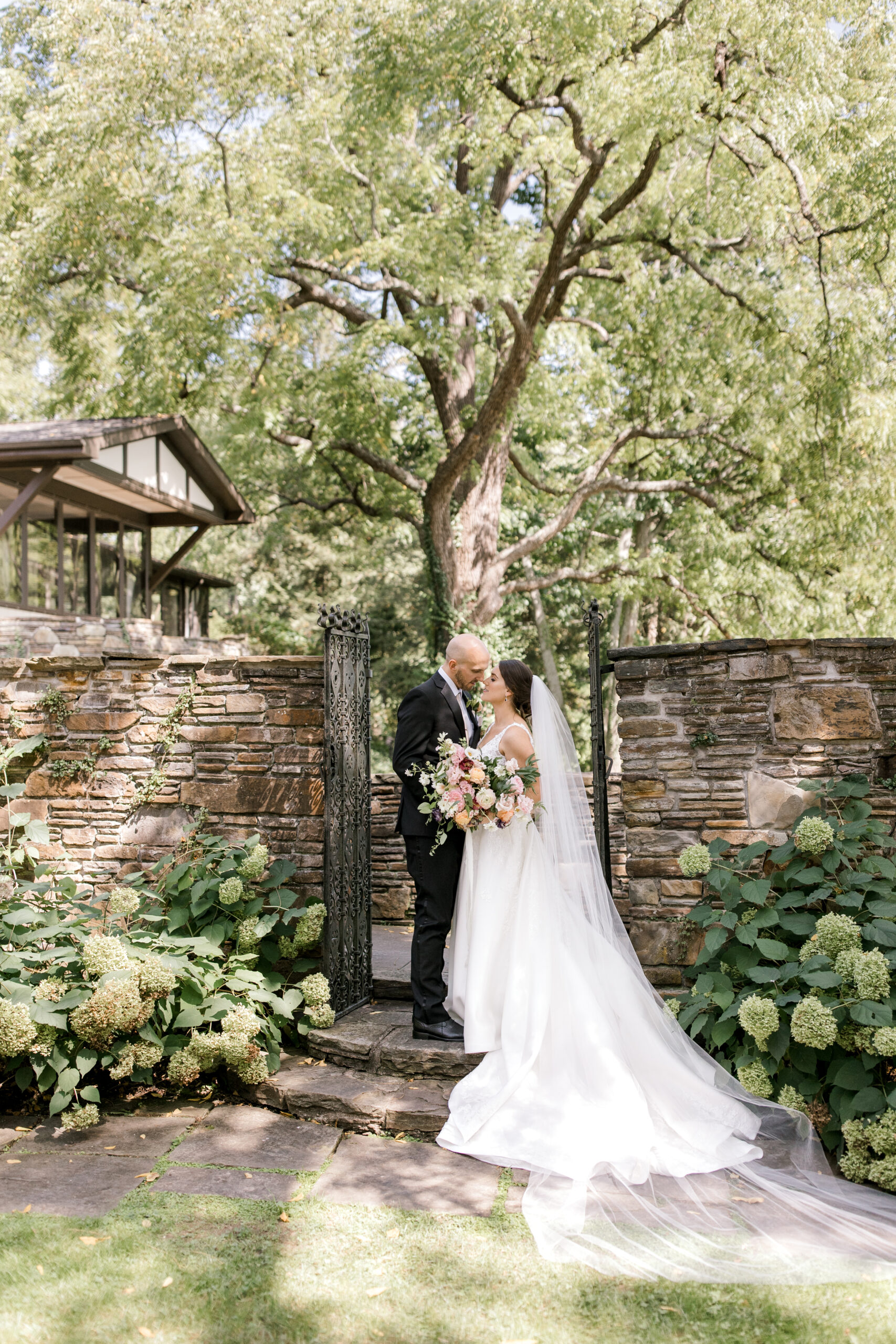 couple leaning against each other about to kiss, standing in an open gateway between stone walls with a tall tree behind them